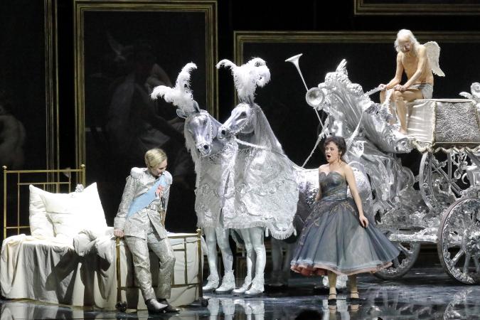 Szene auf einer Theaterbühne: Opulent gekleidete Dame und Herr neben einem Bett, daneben eine Kutsche
