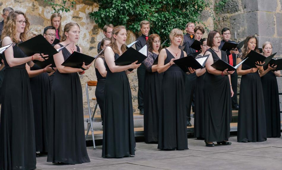 Ein schwarz gekleideter Chor singt im Freien vor einer Mauer aus schwarzen Notenmappen