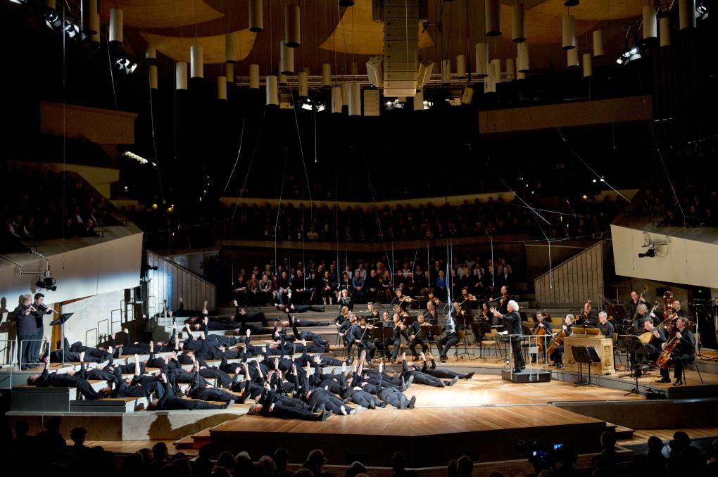 Aufführung der Matthäus-Passion in der Berliner Philharmonie, inszeniert von Peter Sellars. Links liegen schwarz gekleidete Gestalten in mehreren Reihen auf der Bühne. Rechts sind Musikerinnen und Musiker der Berliner Philharmoniker zu erkennen, ebenfalls schwarz gekleidet.  
