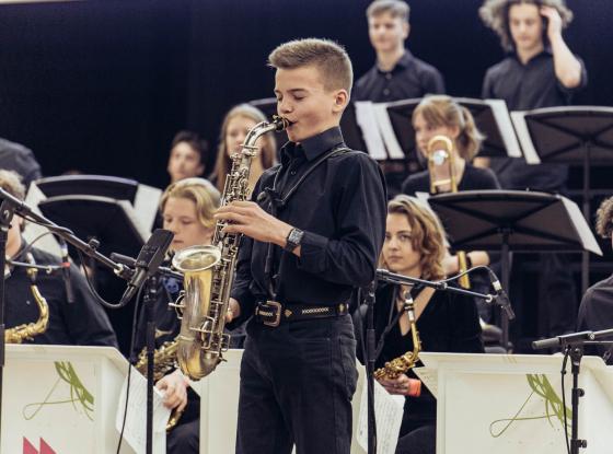 Young saxophonist is playing at a concert, jazz orchestra in the background